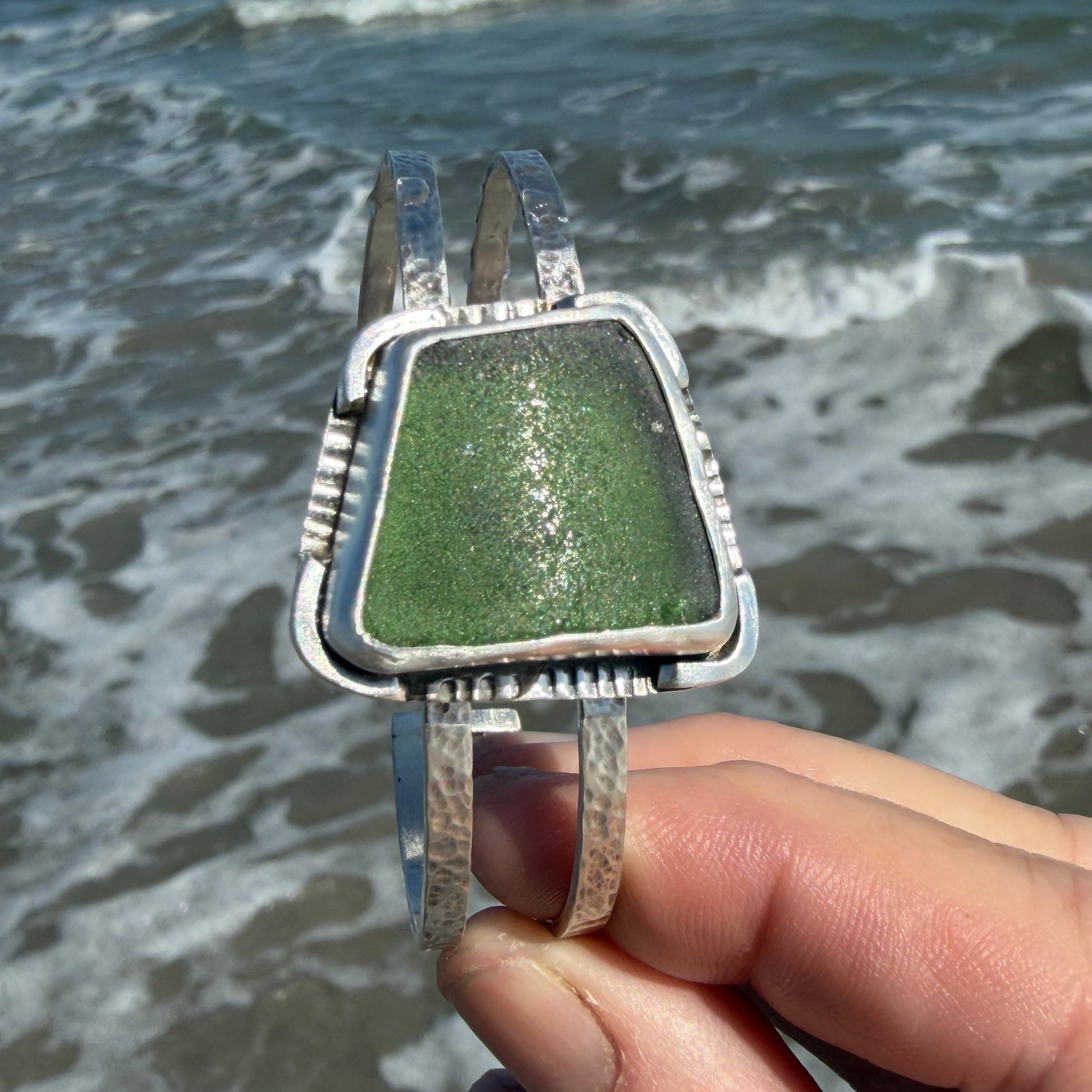 Hand holding a green square sea glass bracelet with silver frame  
 against the ocean 