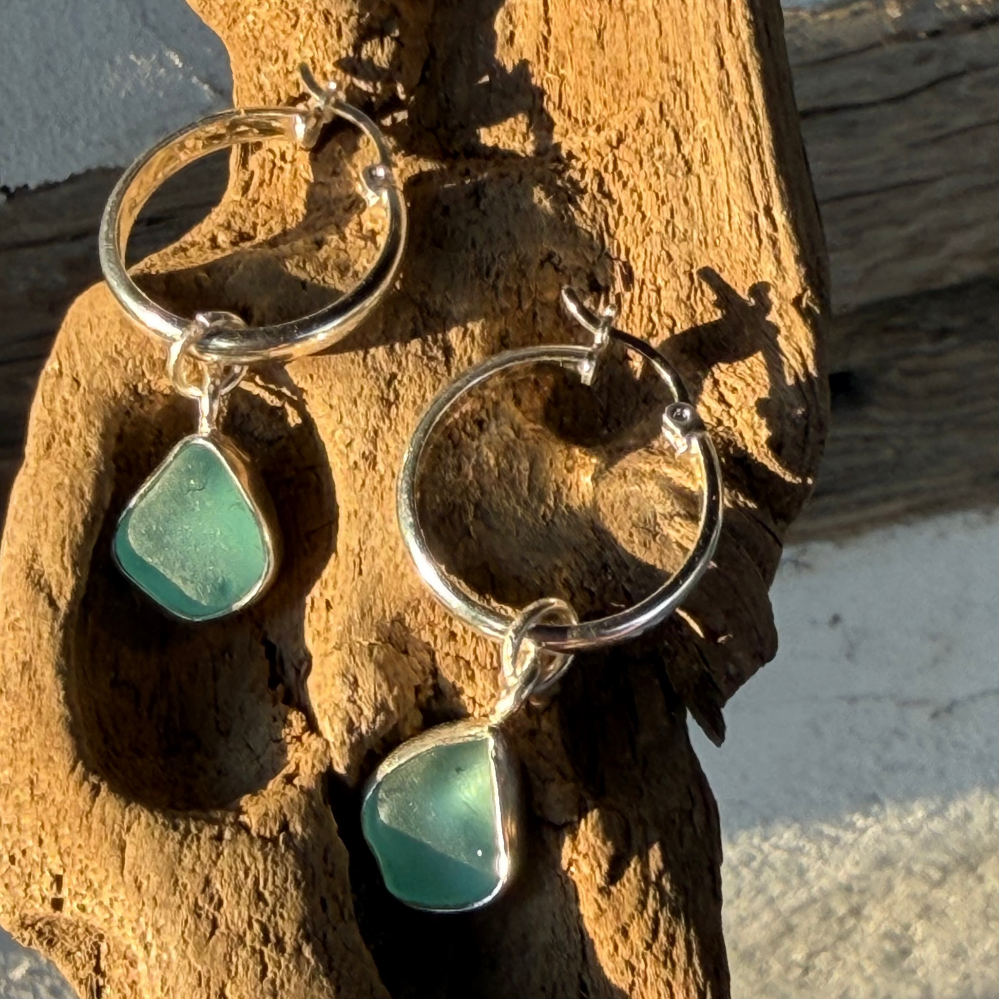 Pair of silver hoop earrings with teal sea glass charms on a wooden surface.
